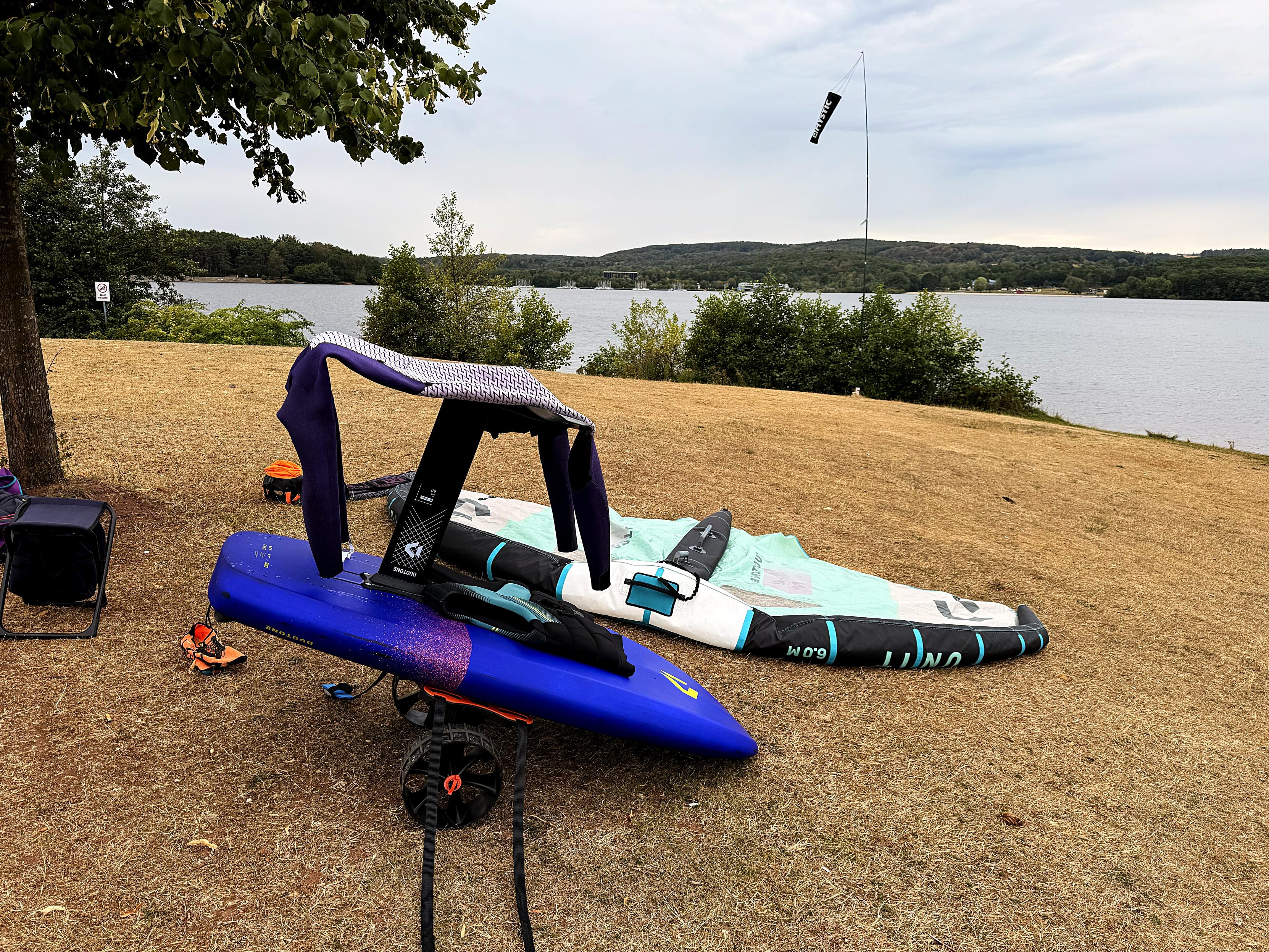 Wing, Foilboard und Neoprenanzug am Seeufer.